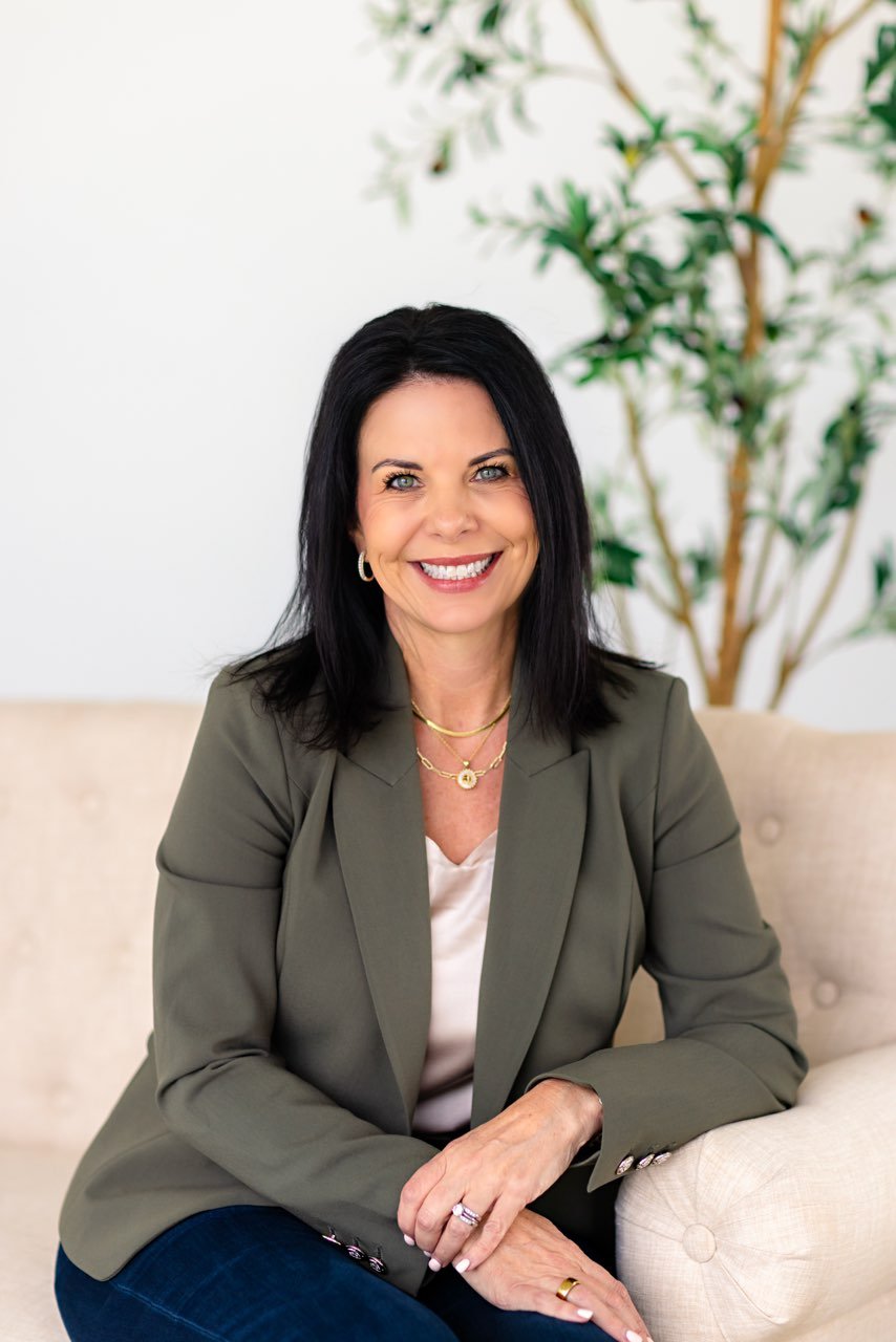 Jodi Ashcraft seated on a sofa in an olive blazer, smiling