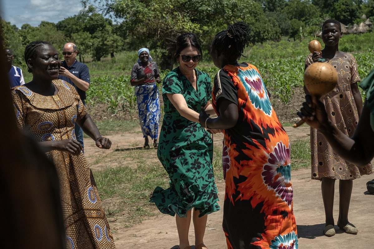 Jodi dancing with women on a dirt road beside a maize field